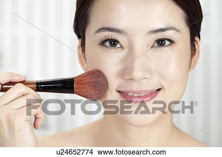 Picture - Beauty Treatment, Close-up of woman holding a blush brush on her face with smile. Fotosearch