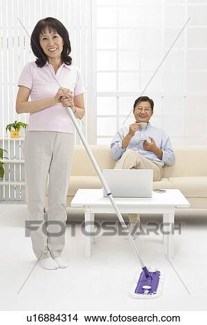 Couple, Wife cleaning the house and husband sitting on sofa and drinking tea View Large Photo Image Picture - Couple, Wife cleaning the house and husband sitting on sofa and drinking tea. Fotosearch