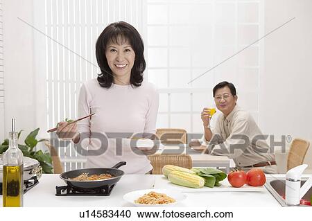 Stock Image - Couple, Wife cooking food and husband drinking near the table with smile. Fotosearch