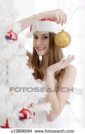 Picture - Young woman wearing Santa hat and holding Christmas bauble with smile. Fotosearch