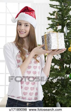 Stock Photo - Young woman wearing Santa hat and holding a wrapped gift with smile. Fotosearch