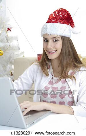 Stock Photo - Young woman wearing Santa hat and using laptop with smile. Fotosearch