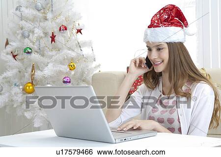 Young woman wearing Santa hat, holding mobile phone and using laptop View Large Photo Image Stock Photograph - Young woman wearing Santa hat, holding mobile phone and using laptop. Fotosearch