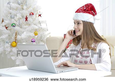 Stock Photograph - Young woman wearing Santa hat, holding mobile phone and using laptop. Fotosearch