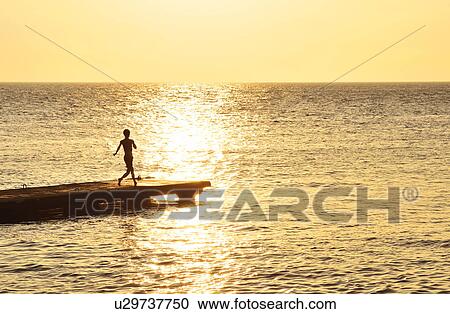Stock Image - Boy running at pier at dusk,. Fotosearch