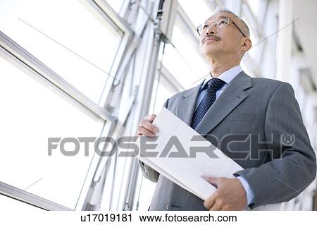 Stock Image - Businessman standing by window, holding blueprint. Fotosearch