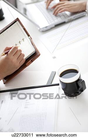 Stock Image - Businesswomen writing in her diary and using laptop. Fotosearch