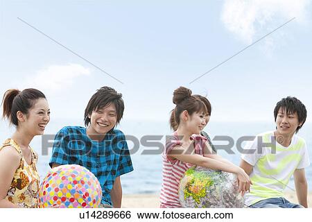 Stock Photograph - Five young people sitting on beach, women holding beach balls. Fotosearch