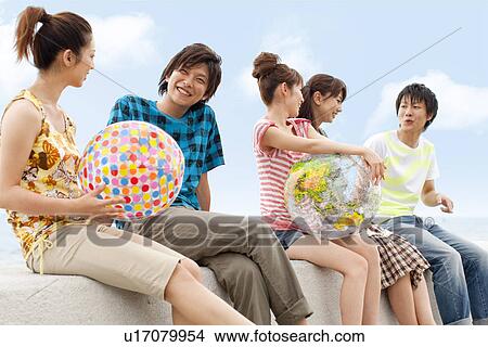 Picture - Five young people sitting on breakwater, women holding beach balls. Fotosearch