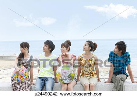 Stock Image - Five young people sitting on breakwater, women holding beach balls. Fotosearch