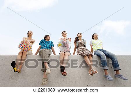 Stock Photo - Five young people sitting on breakwater, women holding beach balls. Fotosearch