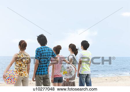Stock Photo - Five young people standing side by side at beach, women holding beach balls. Fotosearch