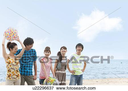Stock Photo - Five young people standing side by side at beach, women holding beach balls. Fotosearch