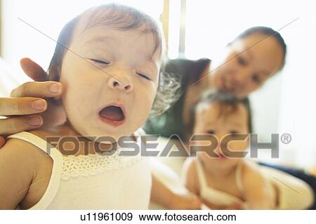 Stock Photo - Mother touching baby girl, twin sister in the background. Fotosearch