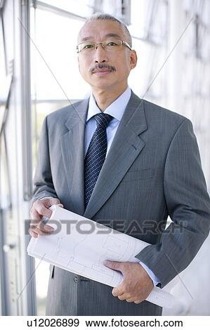 Stock Photo - Portrait of businessman standing by window, holding blueprint. Fotosearch
