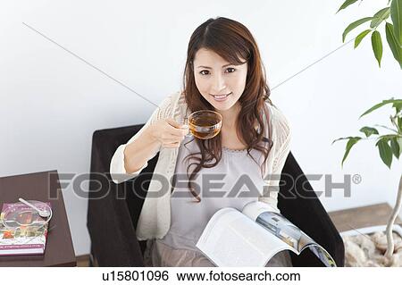 Portrait of young woman sitting on sofa, holding cup of tea and magazine View Large Photo Image Stock Photograph - Portrait of young woman sitting on sofa, holding cup of tea and magazine. Fotosearch