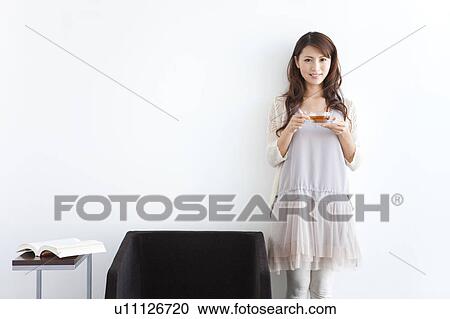 Stock Image - Portrait of young woman standing by sofa, holding cup of tea, white background. Fotosearch
