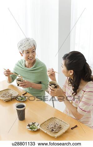 Stock Image - Two women eating soba noodles, smiling. Fotosearch
