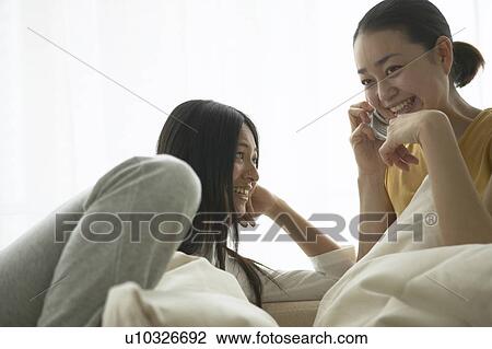 Stock Image - Two women on sofa talking on cell phone. Fotosearch