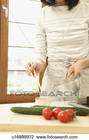 Stock Image - Woman in kitchen, holding pan and chopsticks. Fotosearch