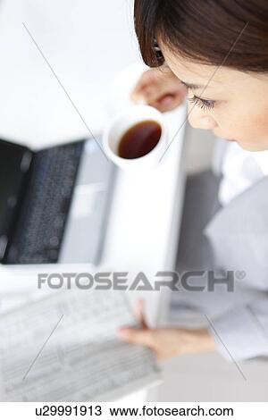 Stock Image - Young businesswoman reading newspaper, holding coffee cup. Fotosearch