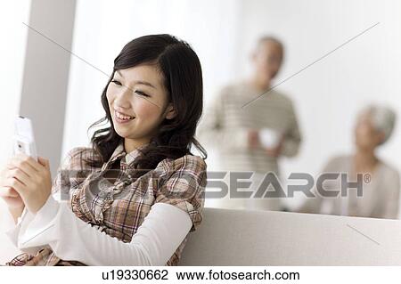 Young woman looking at mobile phone on sofa, parents in background, differential focus View Large Photo Image Stock Image - Young woman looking at mobile phone on sofa, parents in background, differential focus. Fotosearch