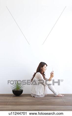 Stock Image - Young woman sitting on floor, using mobile phone, white background. Fotosearch