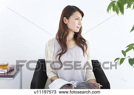 Stock Image - Young woman sitting on sofa, holding magazine, white background. Fotosearch
