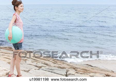 Stock Photo - Young woman standing at beach, holding beach ball. Fotosearch
