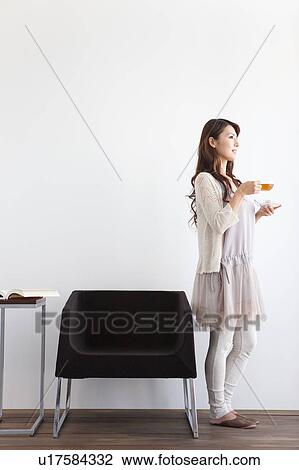 Stock Image - Young woman standing by sofa, holding cup of tea, white background. Fotosearch
