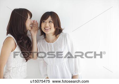 Stock Photography - Young woman whispering friend's ear, white background. Fotosearch