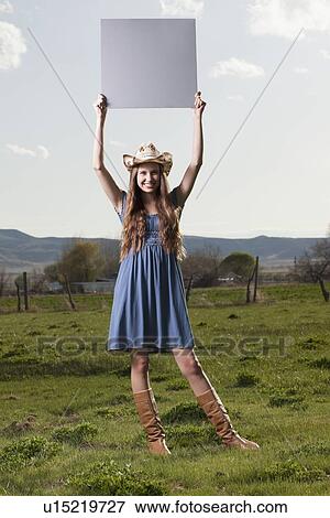 Stock Photo - Cowgirl holding up blank poster while standing in field. Fotosearch