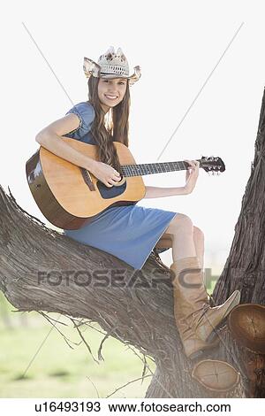 Stock Image - Cowgirl sitting in tree playing guitar. Fotosearch