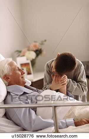 Stock Photograph - Grandfather lying in hospital bed as his grandson holds his hand. Fotosearch