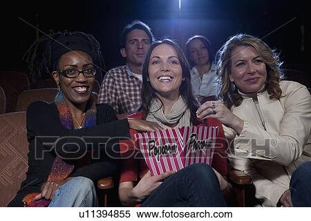 Stock Photography - Three woman sharing popcorn in movie theatre. Fotosearch