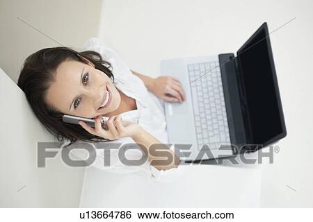 Stock Photograph - Woman talking on phone while working on laptop. Fotosearch