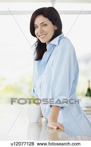 Stock Photo - Brunette woman leaning on kitchen counter. Fotosearch