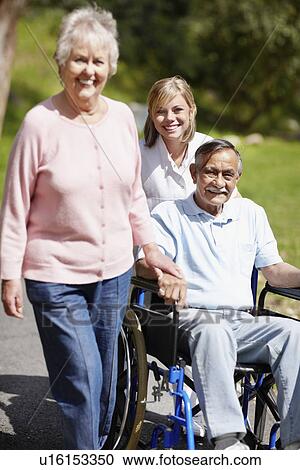 Stock Image - Two women walking with man in a wheelchair. Fotosearch