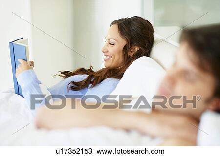Stock Image - Woman reading book in bed beside sleeping husband. Fotosearch