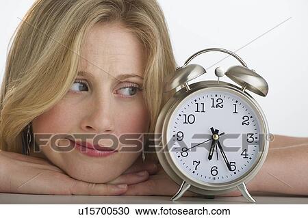 Stock Image - Worried blond woman looking at alarm clock. Fotosearch