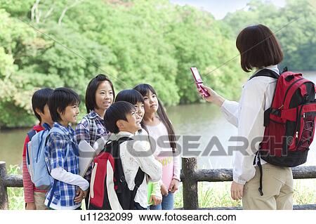 Stock Photo - School children and female teacher in park, teacher taking picture of students by mobile phone. Fotosearch