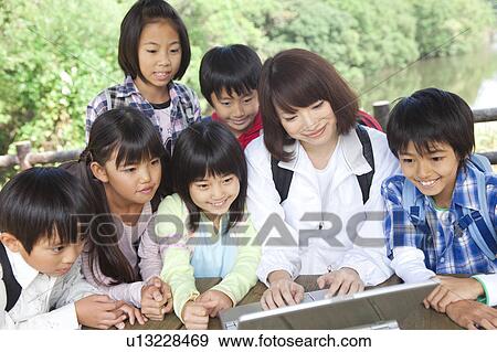 Stock Photo - School children and female teacher using laptop in park. Fotosearch