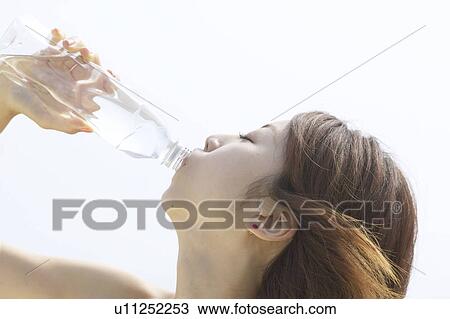 Stock Image - Young woman drinking bottle of water, close up, white background. Fotosearch
