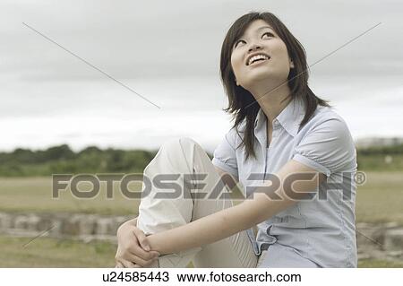 Stock Image - Young woman sitting on bench looking up. Fotosearch