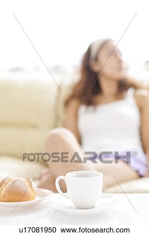 Stock Image - Young woman sitting on sofa, bread and coffee cup on table, differential focus. Fotosearch