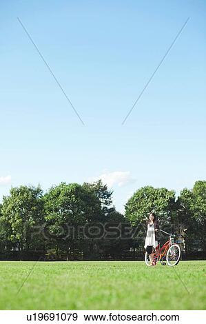 Stock Photo - Young woman standing at a park holding a bicycle, Japan. Fotosearch