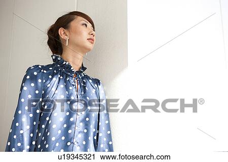 Stock Image - Young woman standing by wall, copy space. Fotosearch