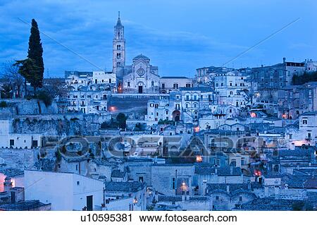 Stock Image - Aerial panorama of ancient town of Matera with tufa houses, Duomo and Campanile on hill beyond, at dusk with night illumination, Puglia, Southern Italy,. Fotosearch