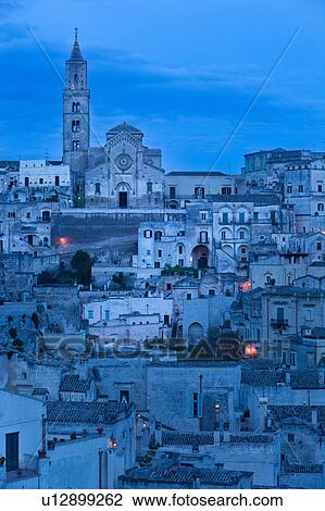 Stock Image - Aerial view of ancient town of Matera with tufa houses, Duomo and Campanile on hill beyond, at dusk with night illumination, Puglia, Southern Italy,. Fotosearch