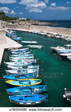 Stock Photography - Aerial view of colourful fishing boats moored in sheltered harbour, vivid green clear water, blue sea and sky beyond, sunny spring afternoon, Salento, Puglia, Southern Italy. Fotosearch